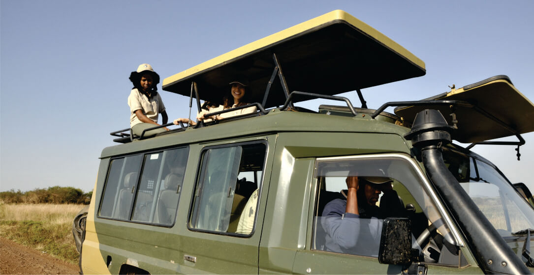 Two women sitting on top of a green open-roofed safari jeep with driver guide in Masai Mara on hired 4x4 safari vehicle