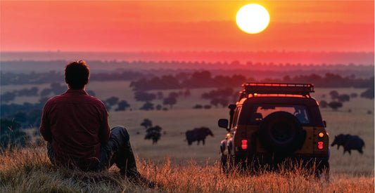 Person sitting on a hill overlooking a sunset with a safari vehicle and elephants in the distance on affordable Masai Mara wildlife safari game drives in Kenya.
