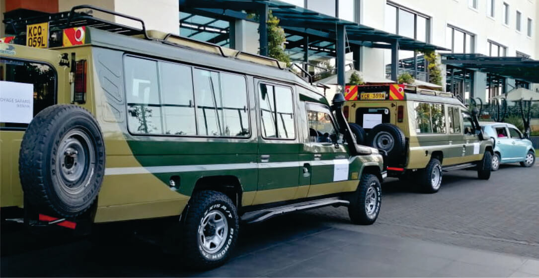 Two green and beige 4x4 jeep safari vehicles parked at a Nairobi city Hotel, awaiting to transfer guests in Kenya