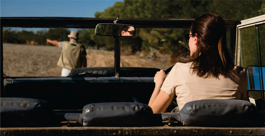 Person sitting in a vehicle looking through binoculars at a field with another person in the background.