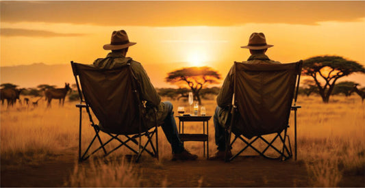 Two people in safari hats sitting in chairs watching a sunset in a savannah.